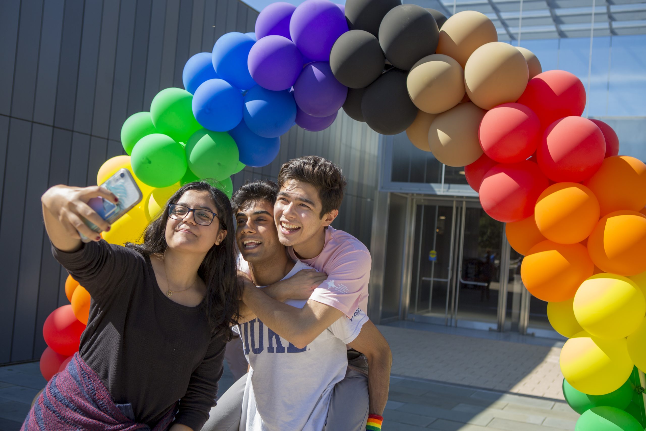 three friends posing for a selfie under a rainbow balloon arch